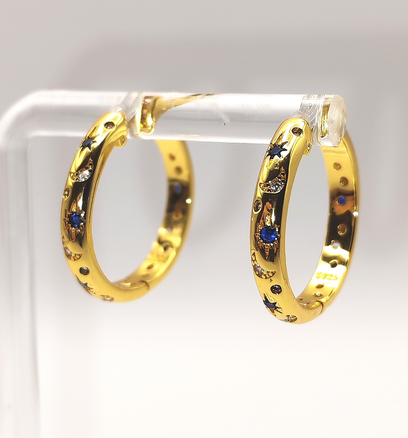 Gold hoop earrings with embedded blue and white stones on a clear stand.