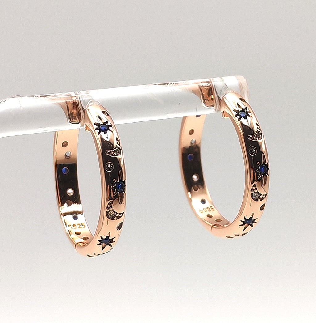 Close-up of rose gold celestial hoop earrings showing blue and white zirconia star and moon detailing.