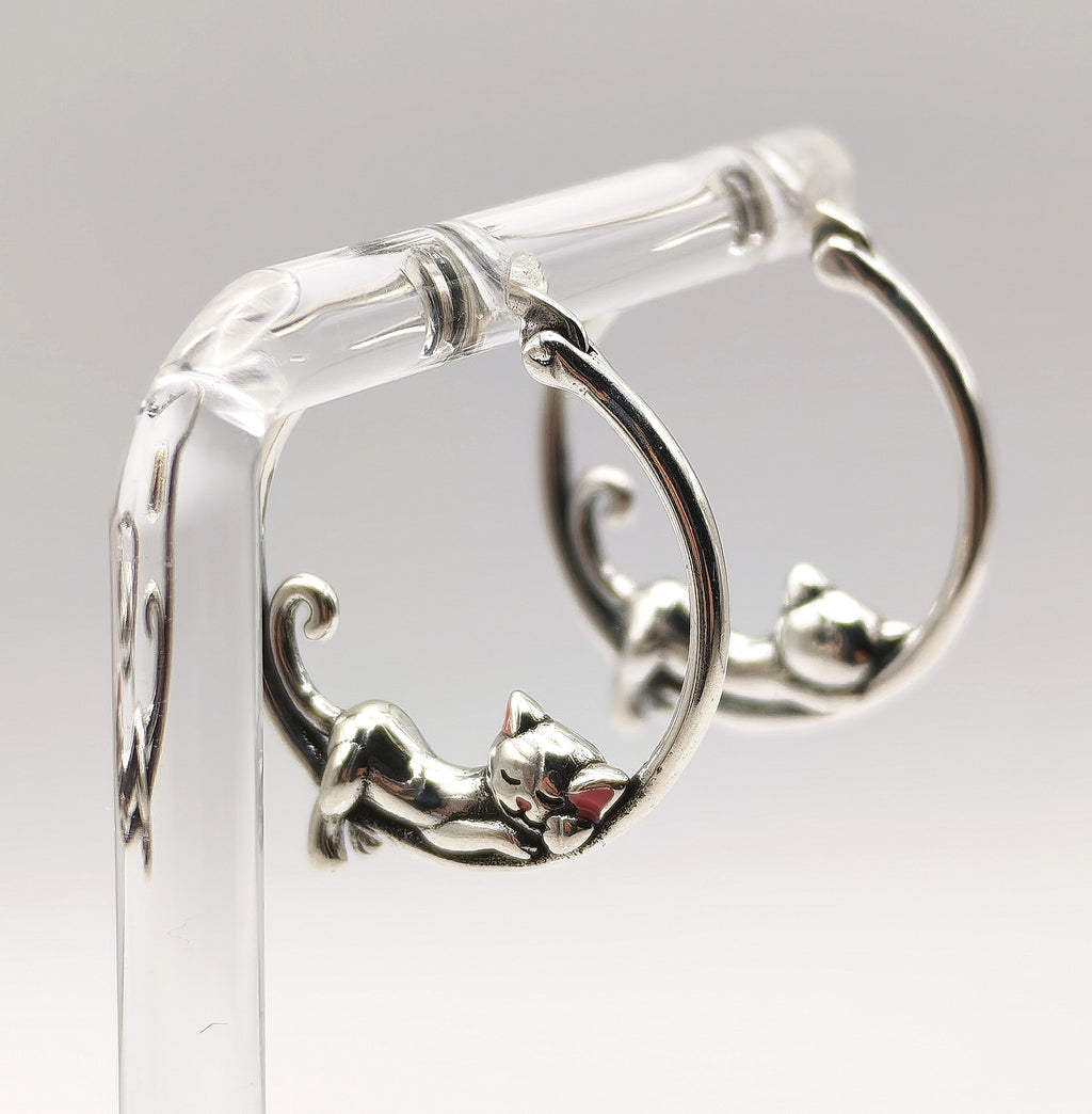 Pair of sterling silver sleeping cat hoop earrings displayed on a clear stand.