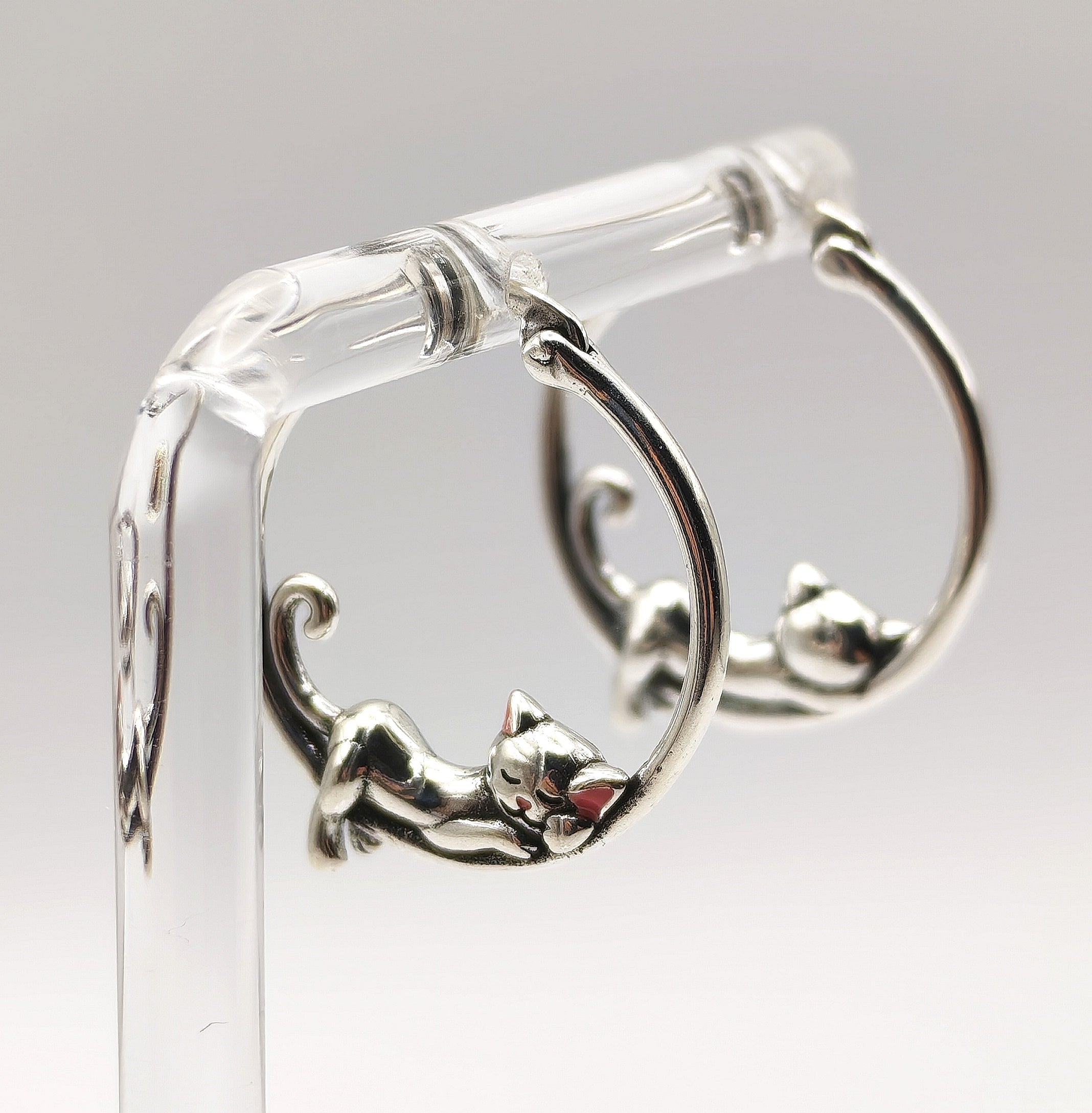 Pair of sterling silver sleeping cat hoop earrings displayed on a clear stand.