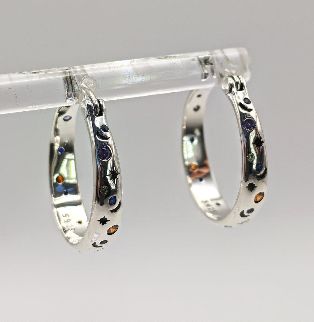 A pair of polished Sterling Silver small hoop earrings featuring an embossed or engraved pattern of crescent moons and stars, with small multi-color CZ accents.