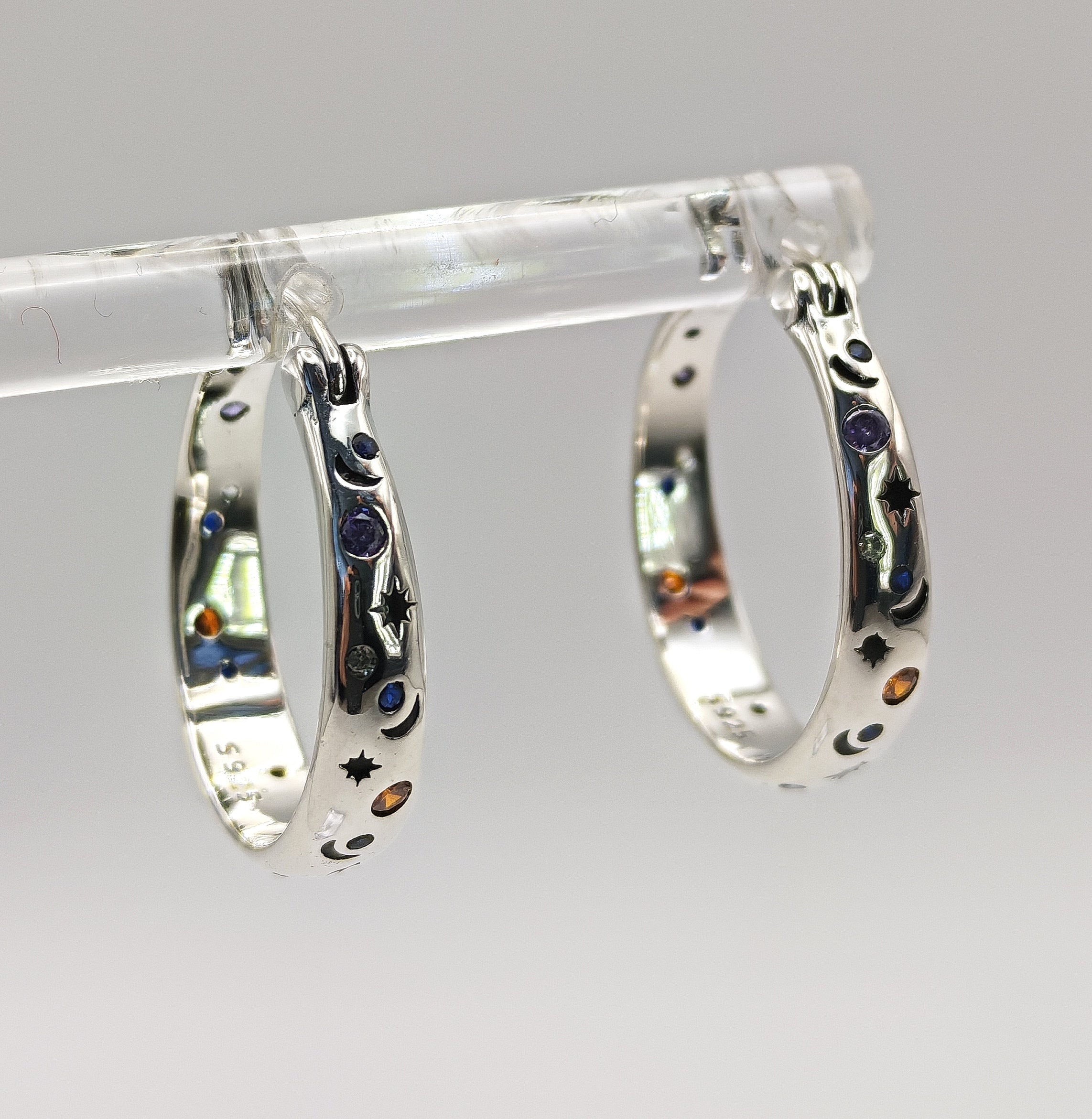A pair of polished Sterling Silver small hoop earrings featuring an embossed or engraved pattern of crescent moons and stars, with small multi-color CZ accents.