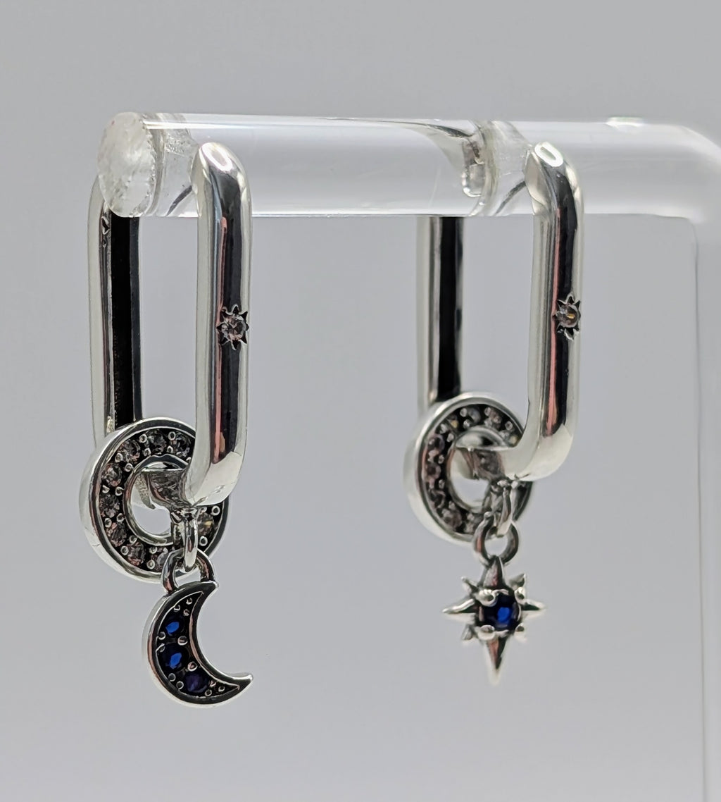 Silver hoop earrings with moon and star designs on a clear stand against a gray background