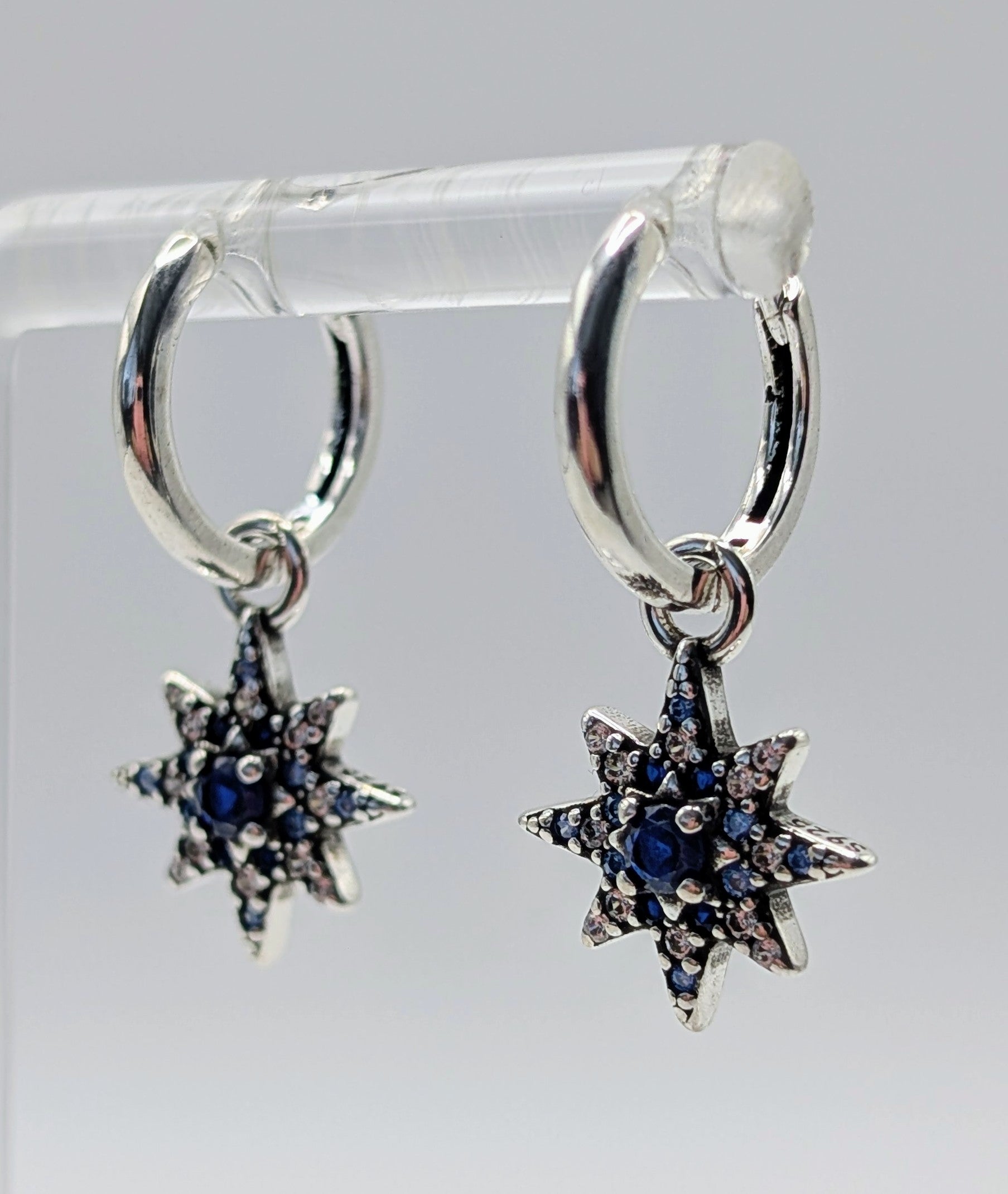 Silver hoop earrings with star-shaped pendants featuring blue stones on a clear stand.